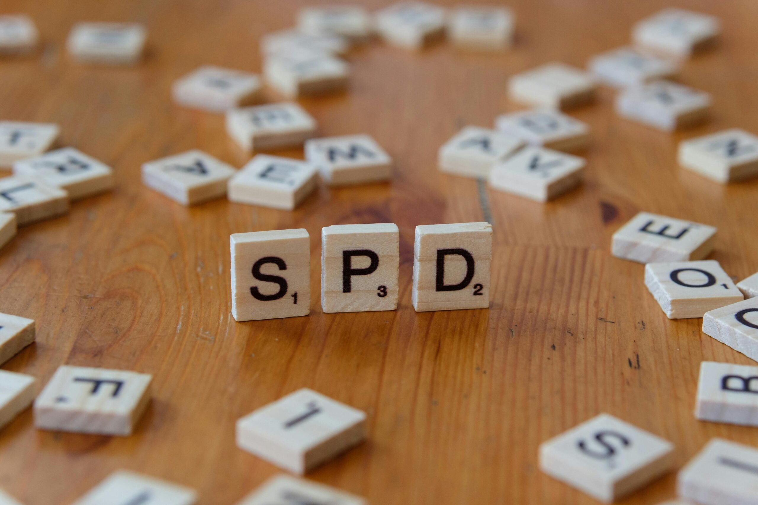 Close-up of 'SPD' spelled with Scrabble tiles on a wooden table, surrounded by scattered letters.